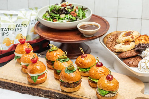 Variety of miniature sliders on brioche on a wooden platter, as a package with cookies, salad, and chips 