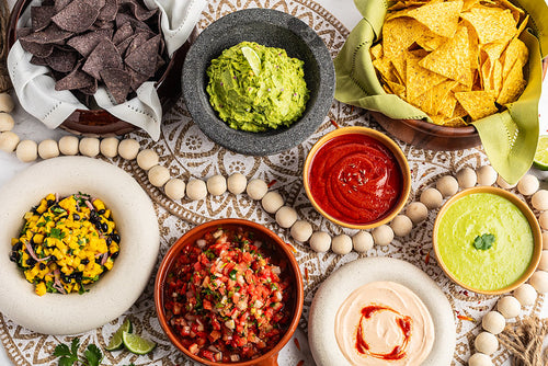 Assorted Mexican snacks including chips, guacamole, salsa, and other dips on a decorative tablecloth.