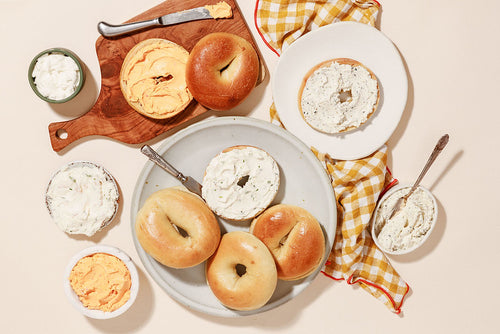 Plain bagels split with a variety of cream cheeses on the side and spread on some halves. Plating is a wood board, a white plate, and some linen cloth