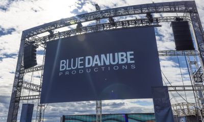 A large outdoor stage screen displays the text 'BLUE DANUBE PRODUCTIONS' in white against a dark blue background, with a metal truss structure and cloudy sky behind it.