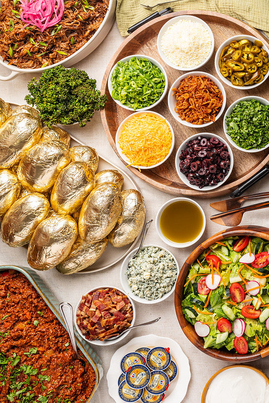 A beautifully styled food spread featuring golden foil-wrapped baked potatoes, a large bowl of fresh green salad with tomatoes, radishes, blue cheese, salsa, pickled onions, shredded cheese, and various side bowls on a light beige tablecloth.