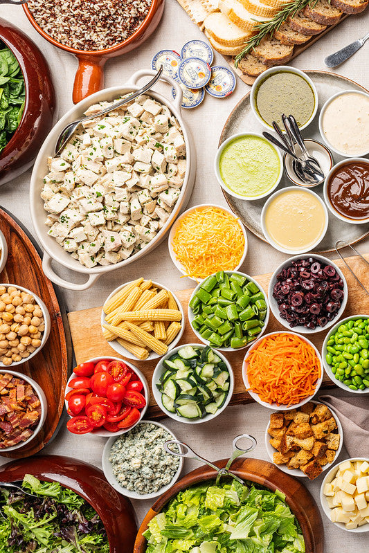 An overhead flat-lay of a catering salad bar, featuring a large bowl of mixed green salad, a large bowl of chicken, bowls of shredded cheese, sliced corn, diced cucumbers, cherry tomatoes, black olives, edamame, blue cheese, and various dressings and toppings on a neutral tabletop.