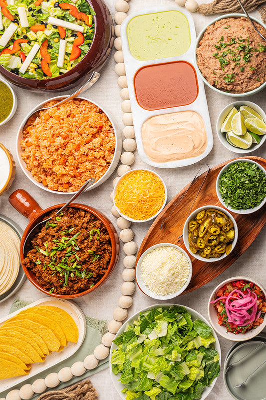 An overhead flat-lay of a Mexican-style taco bar spread with bowls of seasoned rice, seasoned meat, shredded cheese, salsa, lime wedges, lettuce, pickled onions, and a fresh salad. 