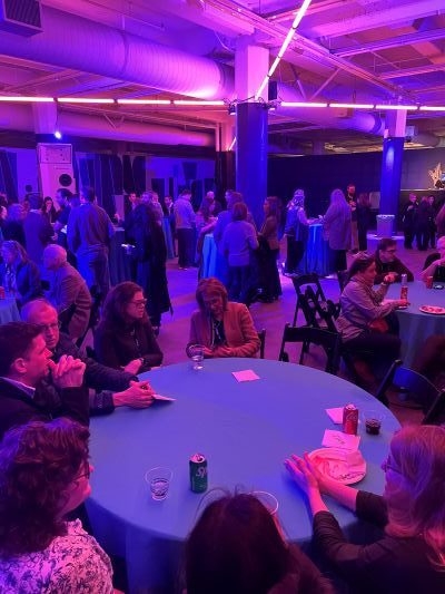 Busy venue with round tables, lots of people both standing and sitting, with strong purple light