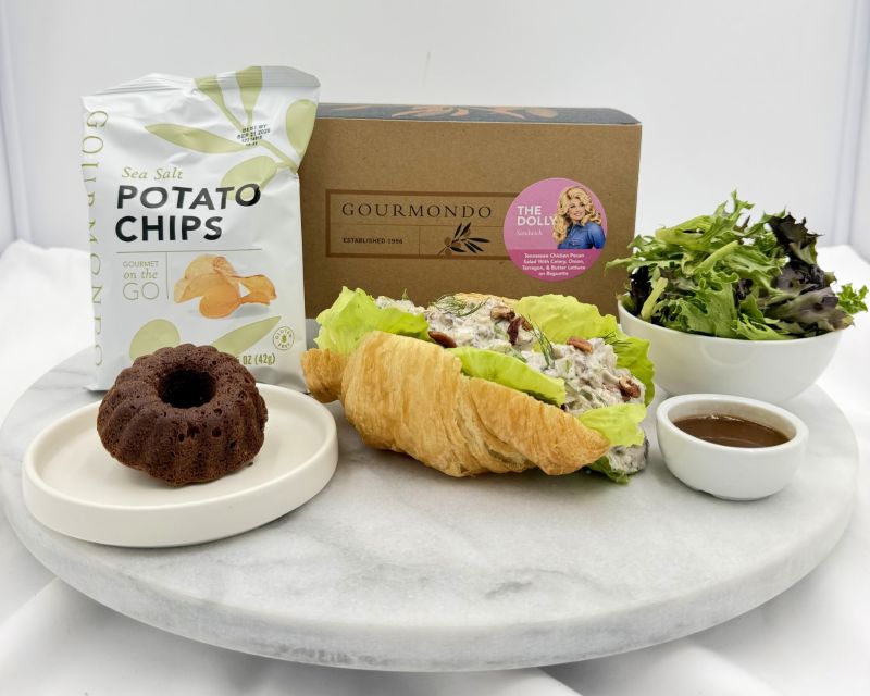 A gourmet food gift set arranged on a round white marble platter against a white studio background. The set includes a bag of Gourmondo sea salt potato chips, a gourmet croissant sandwich with lettuce and cream cheese, a chocolate cake on a small plate, a bowl of mixed greens, and a small bowl of dressing, with a branded box behind the food.