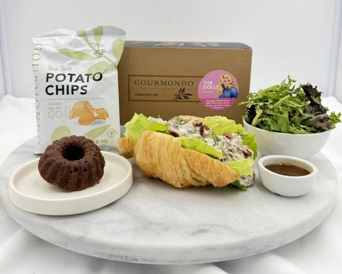 A gourmet food gift set arranged on a round white marble platter against a white studio background. The set includes a bag of Gourmondo sea salt potato chips, a gourmet croissant sandwich with lettuce and cream cheese, a chocolate cake on a small plate, a bowl of mixed greens, and a small bowl of dressing, with a branded box behind the food.
