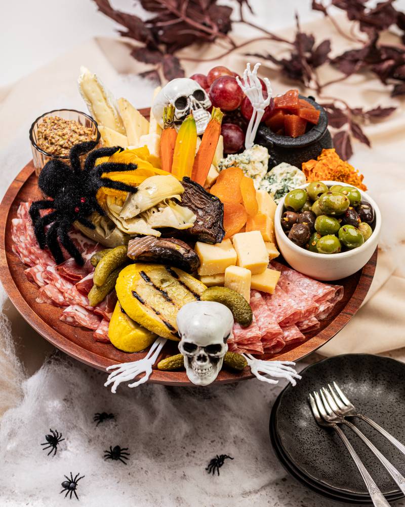 Tray of charcuterie with assorted meats and cheeses, roasted vegetables, olives, and mustard. Halloween decorations of bones and spiders