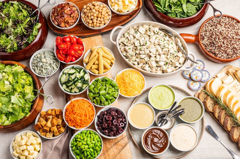 A large, colorful salad bar or picnic spread with various pre-chopped vegetables, dressings, crackers, and bread on a light wood table.