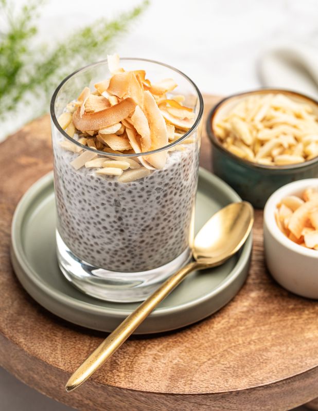 A clear glass cup filled with chia seed pudding topped with shaved coconut, placed on a round sage green saucer with a gold spoon beside it. Two small bowls of coconut sit in the background on a wooden board.