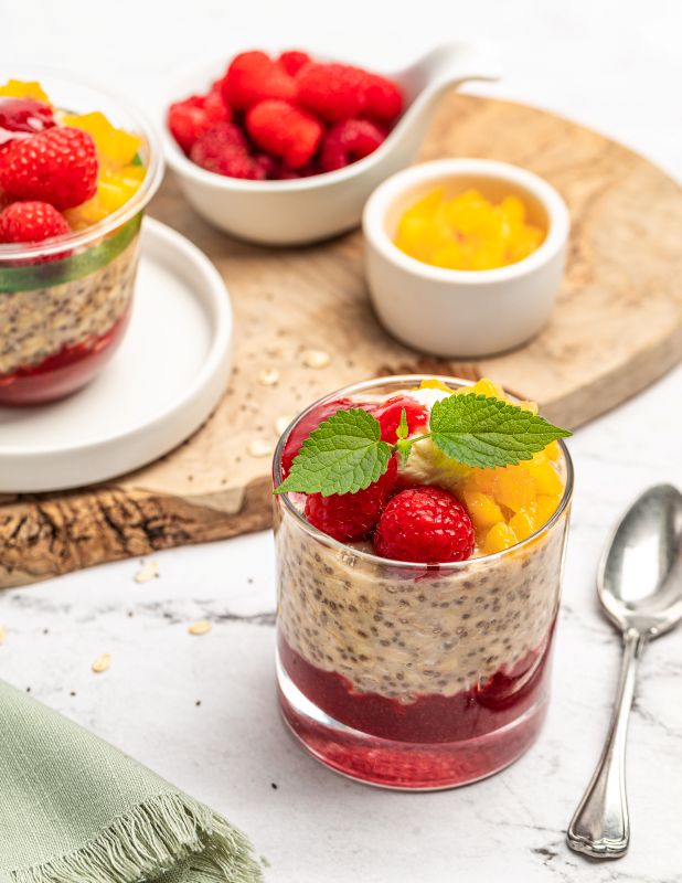 A glass jar of layered chia seed pudding topped with fresh raspberries, yellow fruit, and mint leaves, styled on a marble surface with a spoon, a wooden board, and bowls of fresh berries and fruit in the background.