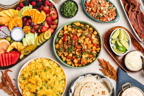 Overhead view of a catered taco bar with bowls of potato hash, scrambled eggs, pico de gallo, wedged limes, cotija, tortillas, and a fruit tray