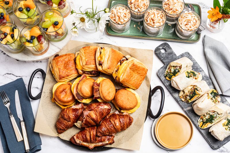 Breakfast catering spread featuring breakfast sandwiches on biscuits and croissants, fresh breakfast wraps, individual fruit cups, chia pudding topped with toasted coconut, and coffee on a marble tabletop.