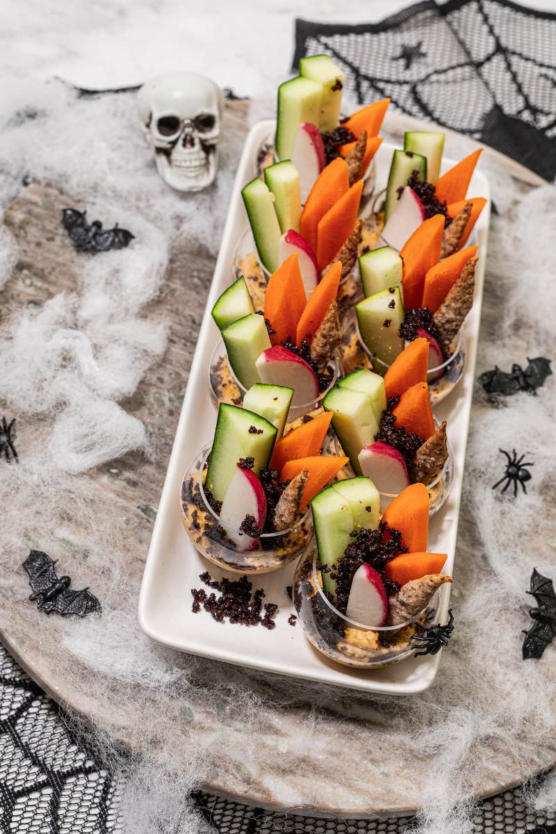 Individual hummus cups with assorted vegetable slices, crushed olives, decorated with Halloween items of skulls and bats