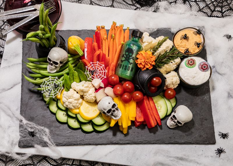 Smoky hummus and tzatziki dips with assorted vegetables on a black tray with Halloween decorations of skulls and spiders