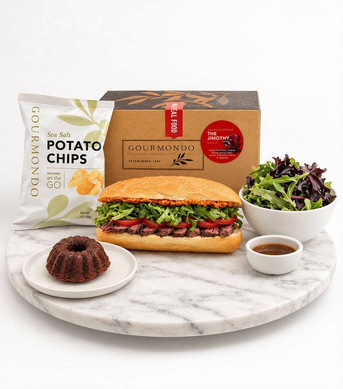 A gourmet food gift set from Gourmondo on a white marble serving board. The set includes a gourmet sub sandwich, a bag of salted potato chips, a chocolate bundt cake, a salad, and a small dipping sauce, arranged in front of a branded brown cardboard box.