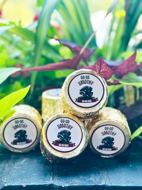 Four small cakes wrapped in shiny gold foil, each topped with a round white label reading 'GO-GO JIMOTHY' with a black animal graphic and smaller text, arranged on a dark blue surface with blurred green and burgundy leaves in the background.