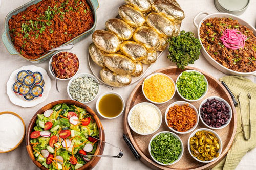 An overhead flat-lay of a large, colorful charcuterie or picnic spread on a light beige tablecloth. The spread features a large loaf of golden-brown bread rolls, a bowl of fresh green salad with tomatoes and radishes, a tray of shredded orange and white cheese, various small bowls of sliced vegetables, pickles, and blue cheese, a large serving of red meat chili or salsa, and a serving of shredded meat with pickled onions.