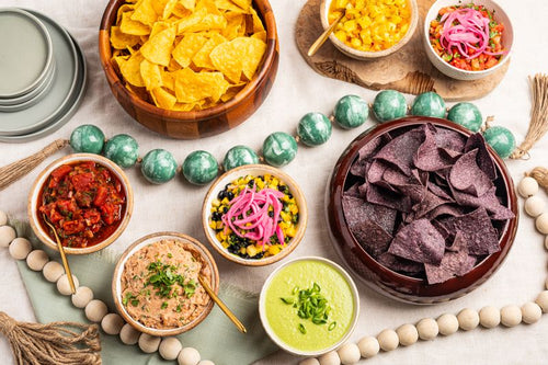 An overhead styled flat-lay of Mexican-style tortilla chips and various colorful dips in small bowls, arranged with decorative beads and plates on a light fabric background.