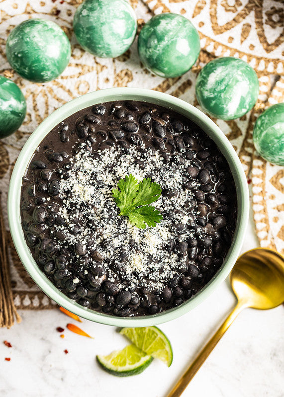 Dish with black beans, topped with cotija and cilantro leaf. Adorned with lime wedges and traditional Mexican decorations