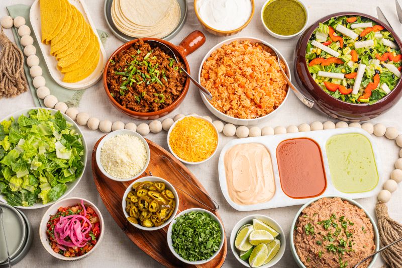 A beautifully arranged Mexican food spread featuring tortillas, seasoned rice, shredded beef, fresh lettuce, pickled onions, jalapeños, salsa, guacamole, lime wedges, and hummus, styled with wooden beads on a light surface.
