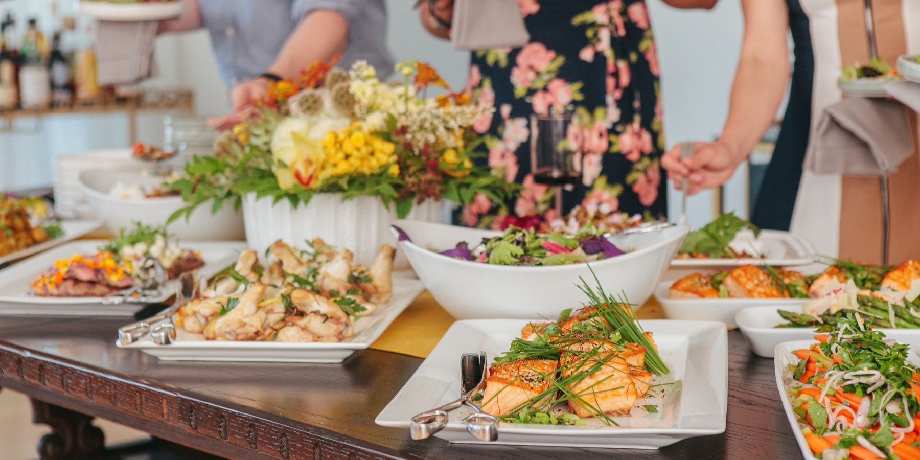 Buffet of assorted proteins and sides with floral centerpiece, with people plating up in the background